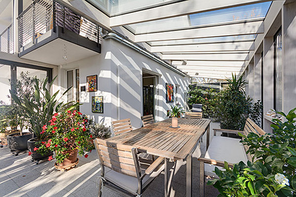 Moderne überdachte Terrasse mit Holztisch, Stühlen und vielen grünen Pflanzen.