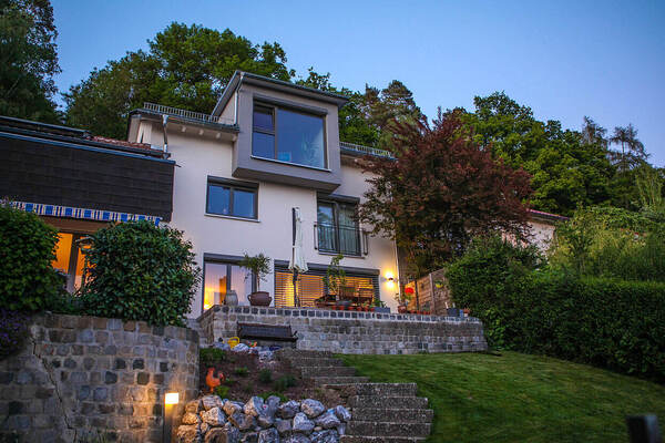 Modernes Einfamilienhaus mit großer Terrasse und Garten am Waldrand bei Sonnenuntergang.
